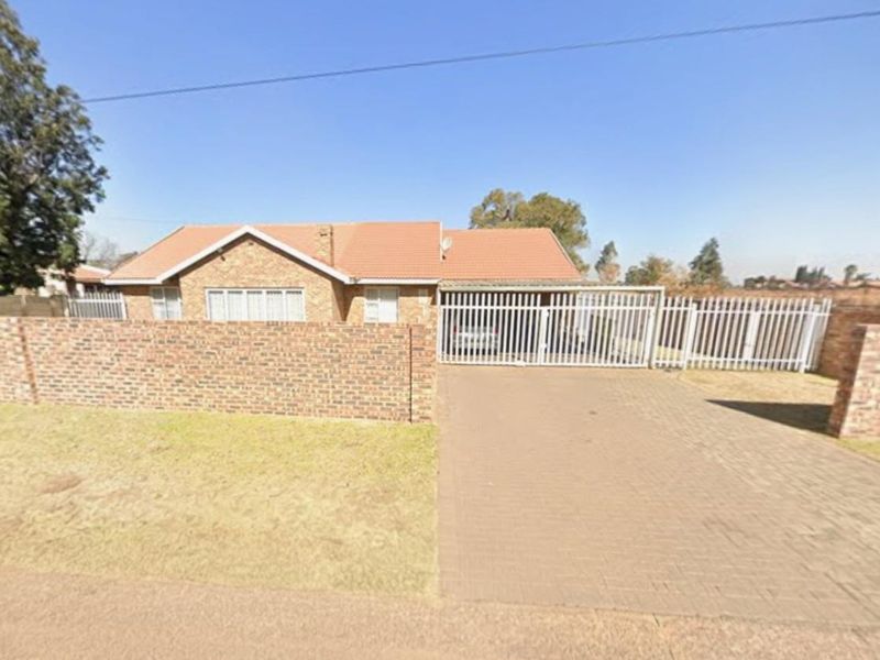 A single-story brick house featuring a tiled roof, paved driveway, and secure perimeter walling with a metal gate.