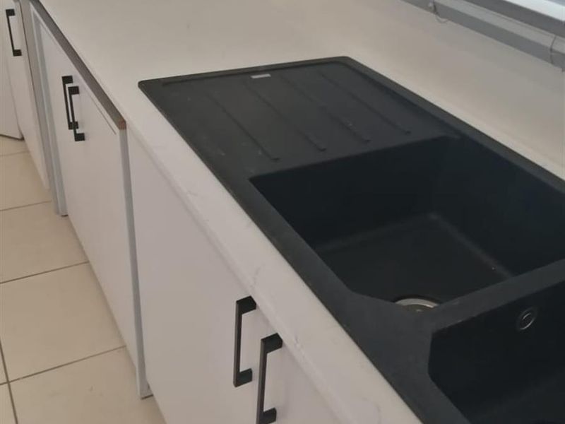 A modern kitchen area featuring white cabinetry, a contrasting black composite sink, and light-colored floor tiles.