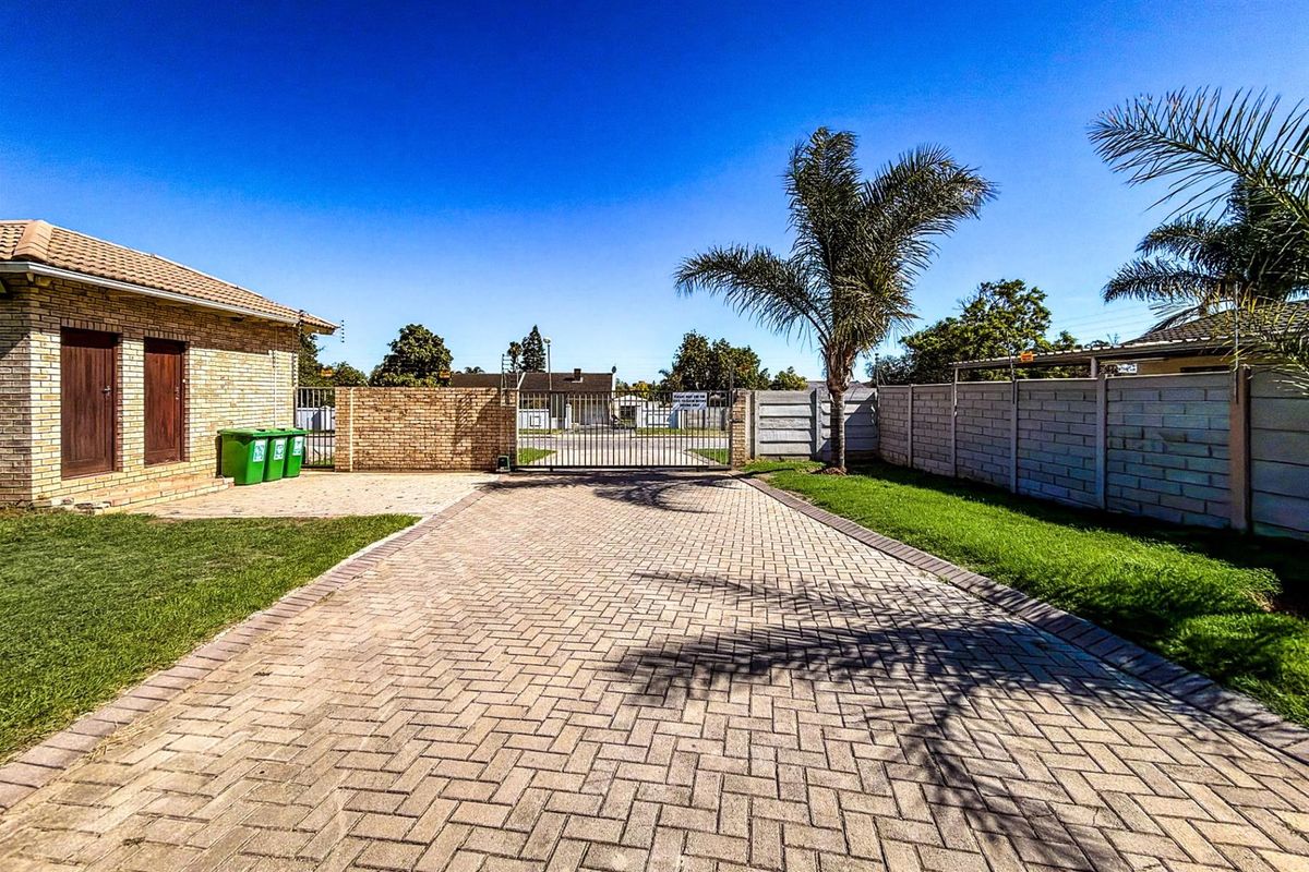 A well-maintained residential property entrance featuring a paved driveway, secure metal gate, brick boundary walls, and established lawn area.