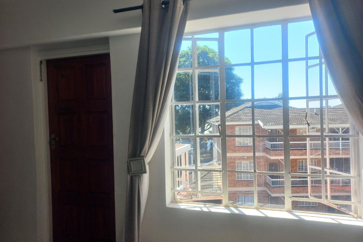 A bright bedroom featuring a large window with security bars, offering a view of the surrounding apartment complex, and a solid timber door.