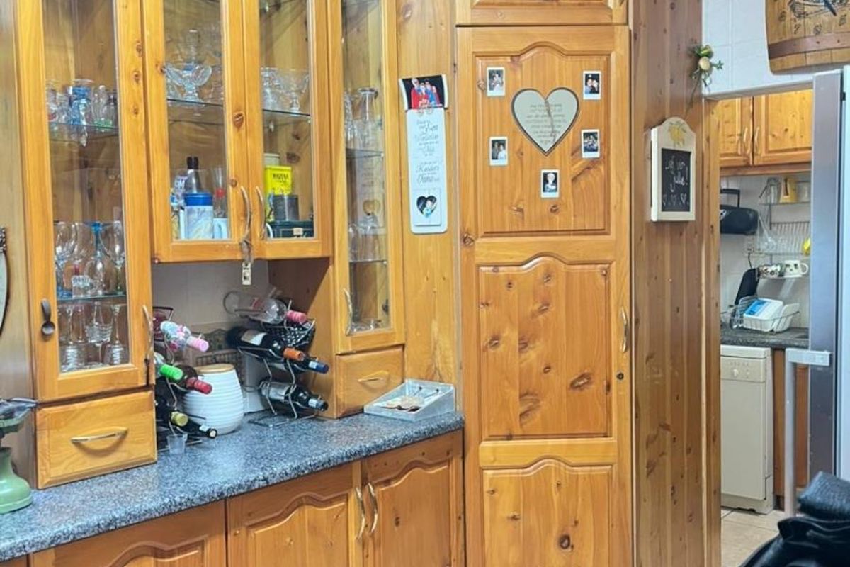 A traditional kitchen featuring extensive solid wood cabinetry, glass-front display cupboards, and granite-look laminate countertops, with an adjacent scullery area visible.