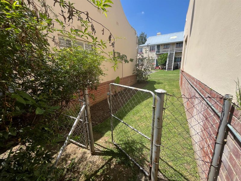 A side garden pathway featuring a chain-link gate, brick boundary walls, and a grassed area leading towards the rear of the townhouse complex.