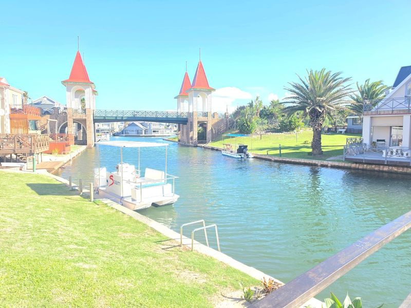 A scenic view of a canal-side residential area featuring a decorative bridge with towers, landscaped lawns, and private water access.