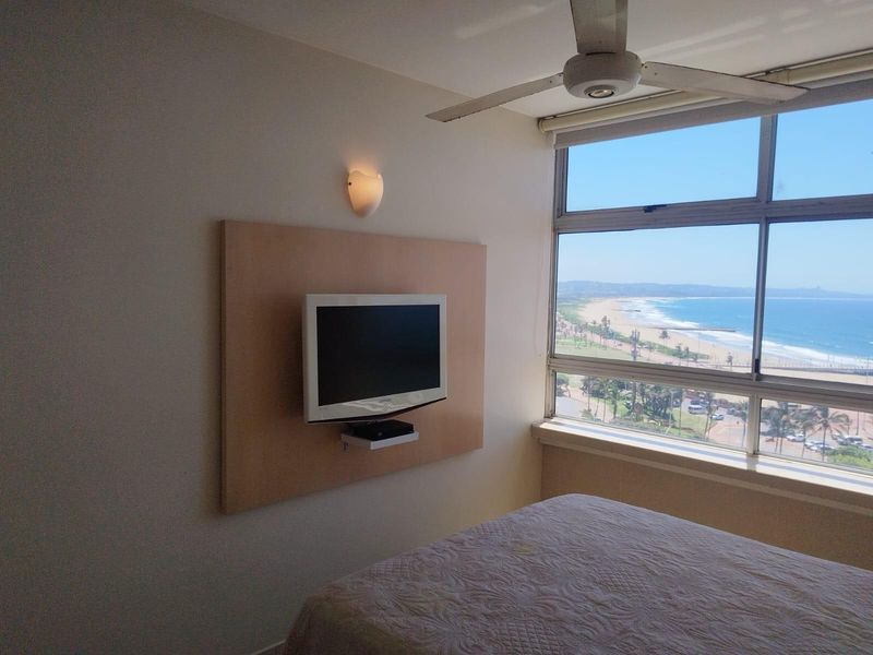 Bedroom featuring a large window with panoramic ocean and beach views, equipped with a ceiling fan and wall-mounted media panel.