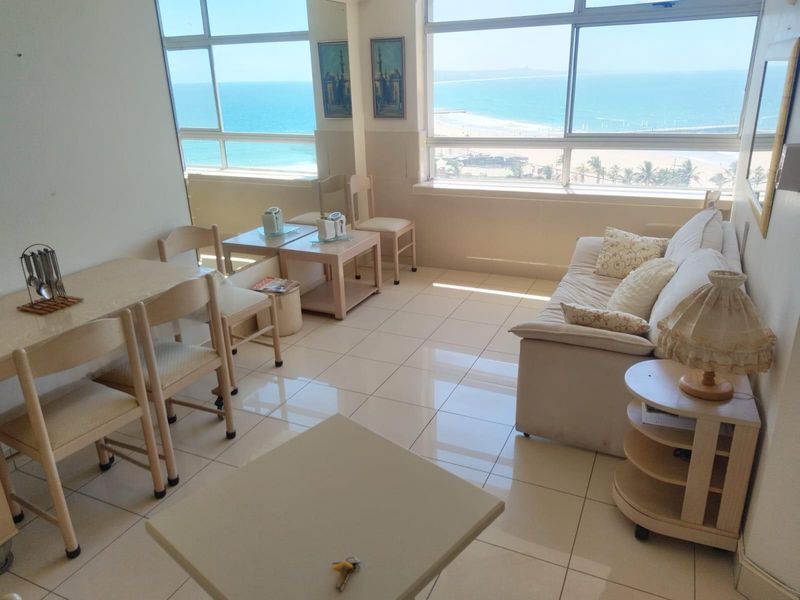 Bright open-plan living and dining area featuring large windows with expansive ocean and beach views, tiled flooring, and natural light.