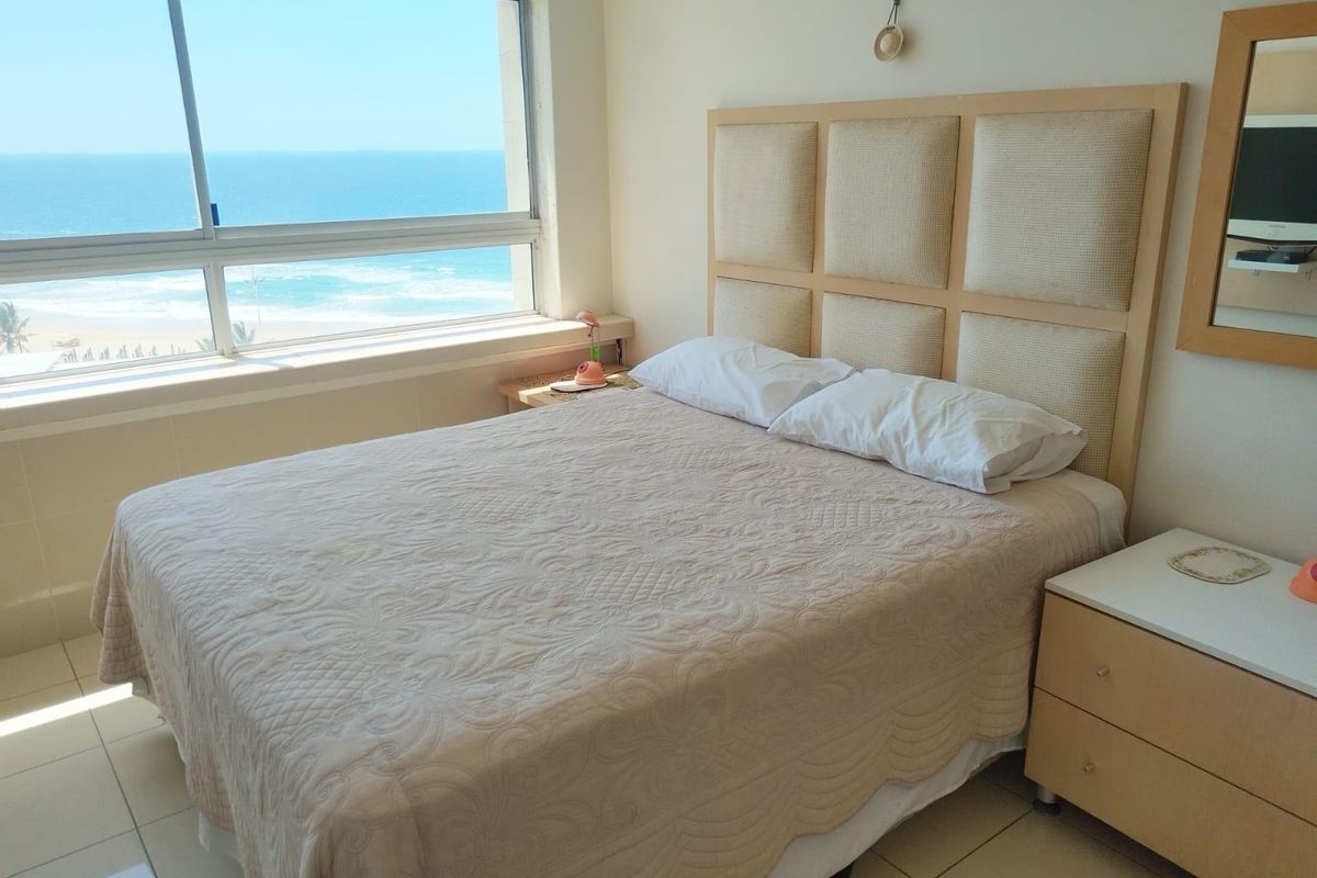 A bedroom featuring large windows with direct ocean views, tiled flooring, and a neutral color palette.