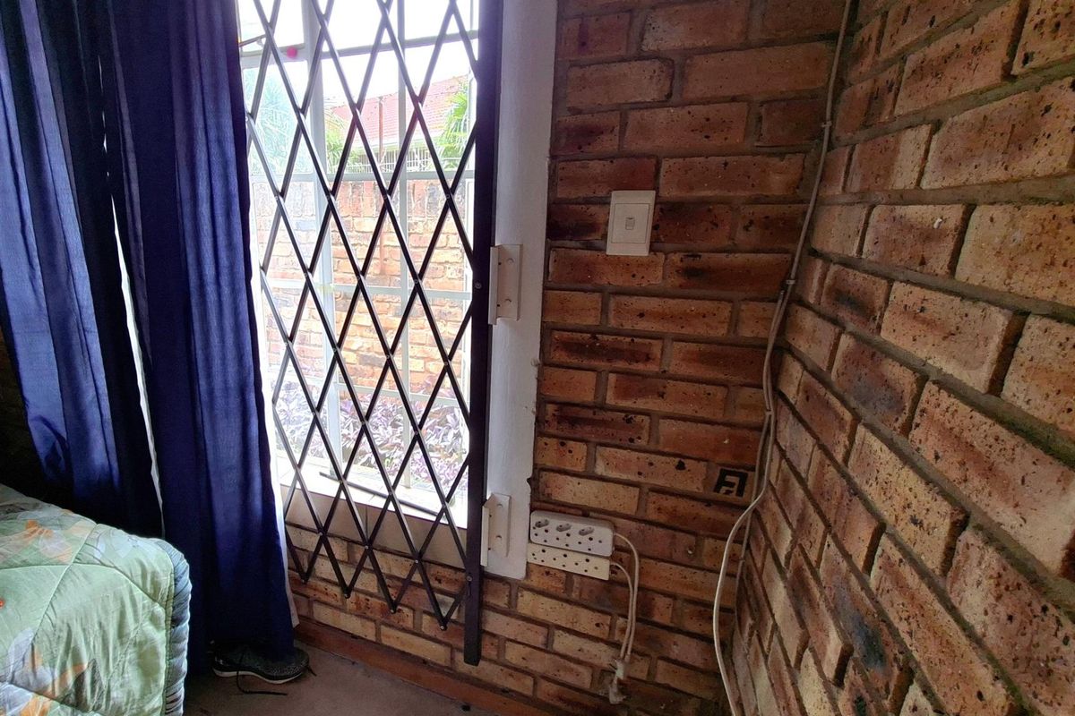 A bedroom corner featuring exposed brick walls, a security gate on the window, and visible electrical wiring.