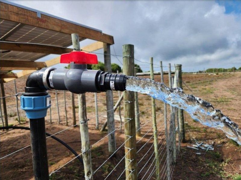 A functional water irrigation tap and pipe system installed on the farm property, featuring a ball valve and solar panel structure in the background.
