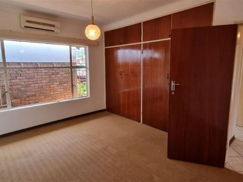 A spacious, empty bedroom featuring built-in wooden cupboards, carpeted flooring, and an air conditioning unit, offering a blank canvas for potential updates.