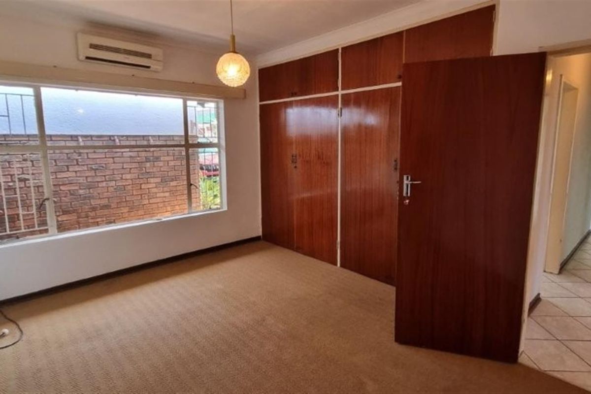 A spacious, empty bedroom featuring built-in wooden cupboards, carpeted flooring, and an air conditioning unit, offering a blank canvas for potential updates.