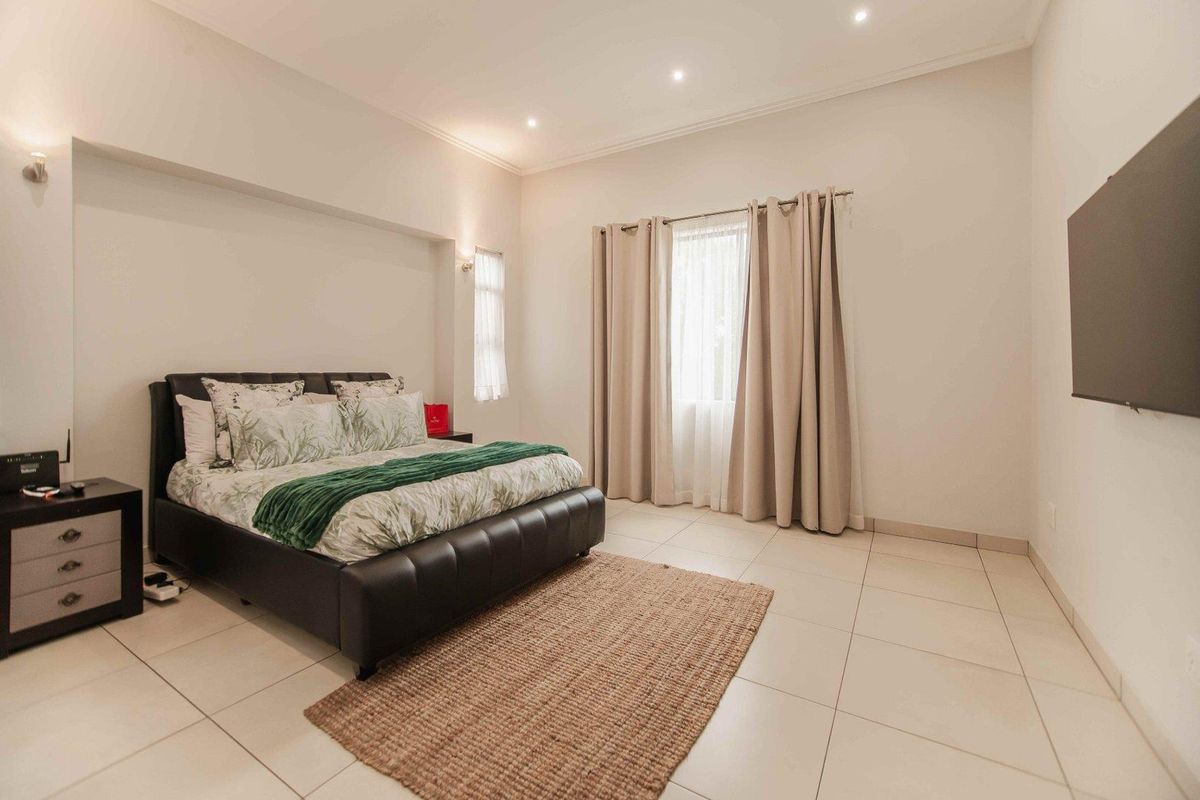 A spacious, well-lit bedroom featuring neutral tiled flooring, recessed ceiling lighting, and a wall-mounted television, offering a clean and modern living space.