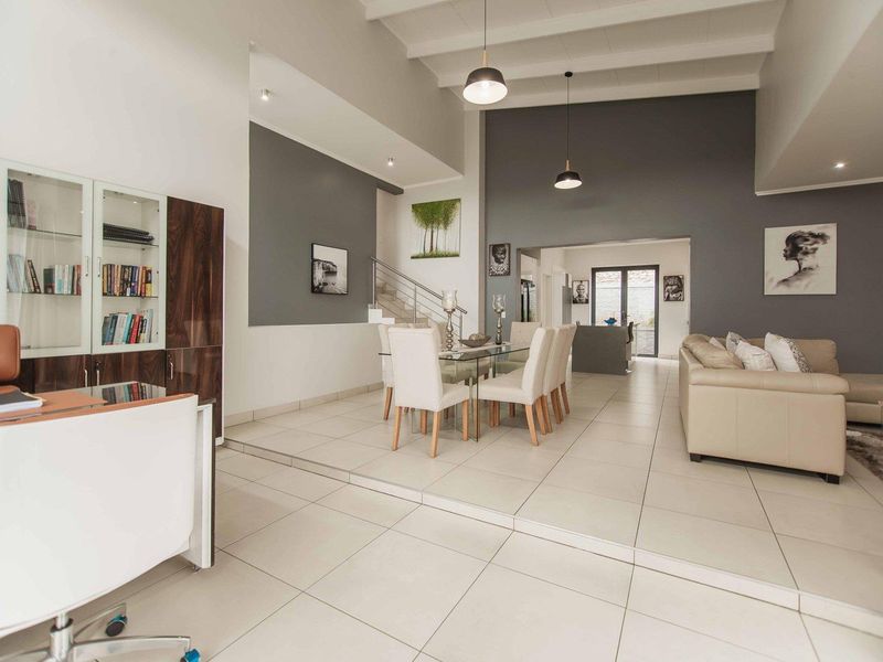 Spacious open-plan living and dining area featuring high ceilings with exposed beams, modern tiled flooring, and a staircase with a contemporary metal railing.