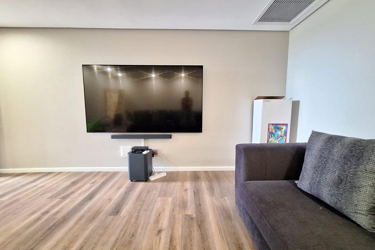 A modern living room featuring light wood-look flooring, neutral wall tones, and a wall-mounted television with integrated ceiling air conditioning vents.