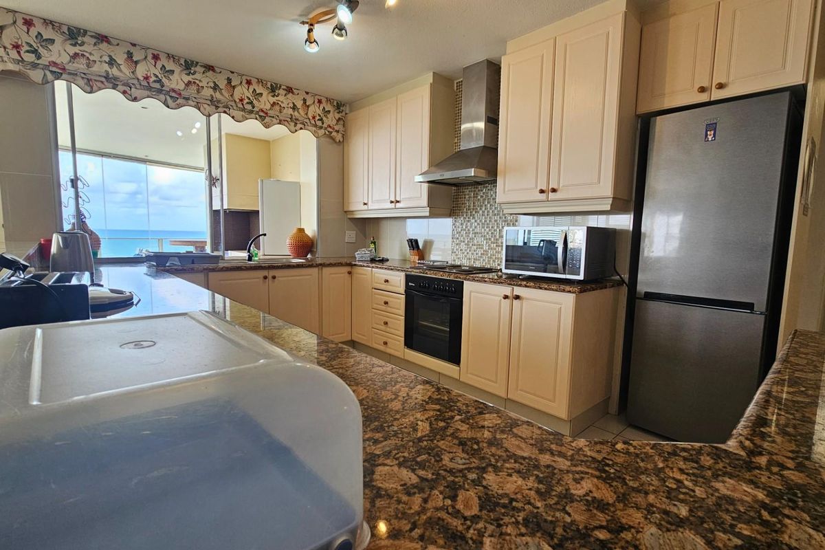 A functional kitchen featuring granite countertops, built-in oven, extractor fan, and light-toned cabinetry, with an open-plan layout overlooking the ocean.