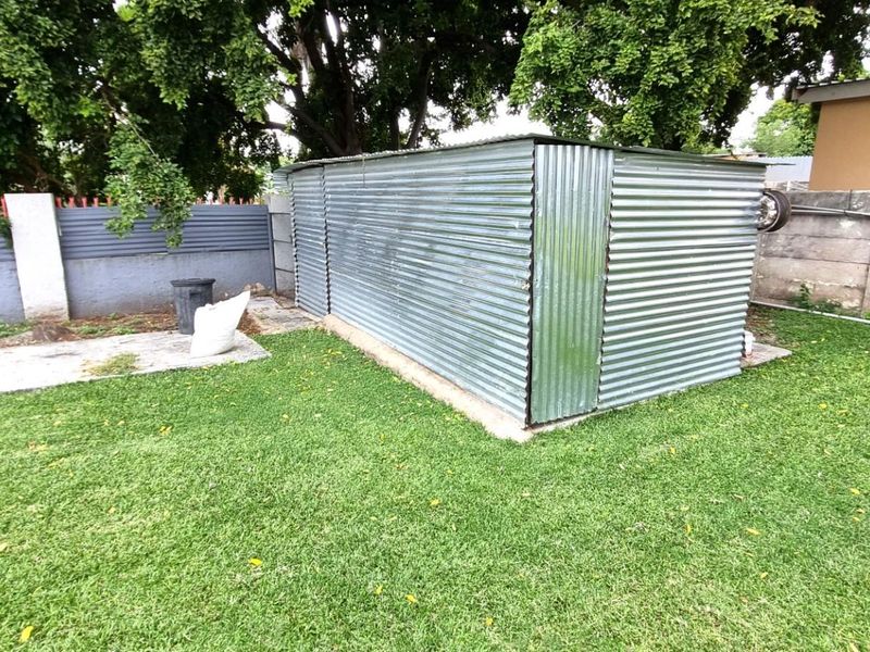 A backyard area featuring a corrugated metal utility shed set on a concrete base, surrounded by a lawn and perimeter boundary wall.