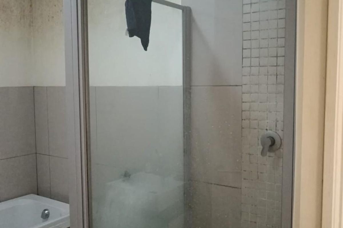A bathroom featuring a glass shower enclosure, tiled walls, and a bathtub, currently showing significant signs of wear and surface deterioration.