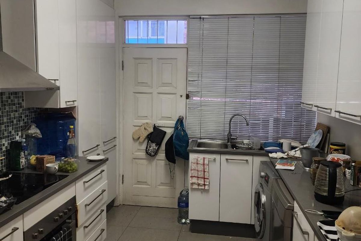 A functional galley-style kitchen featuring white cabinetry, integrated oven and hob, stainless steel sink, and tiled flooring with direct access to an exterior door.