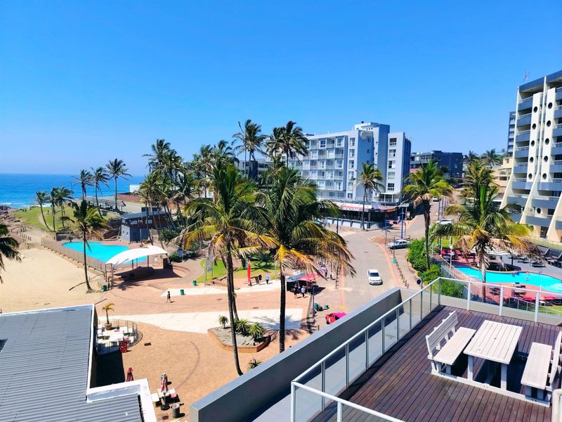 Elevated view overlooking the coastal area, featuring a balcony with decking, nearby swimming pools, palm trees, and proximity to the beach.