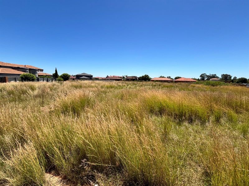 A wide-angle view of a vacant residential plot covered in tall grass, situated within an established suburban neighborhood.