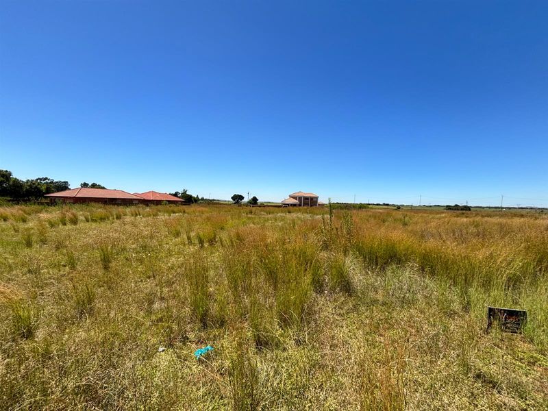 A wide-angle view of a vacant residential plot featuring natural grassland with surrounding neighborhood properties visible in the distance.
