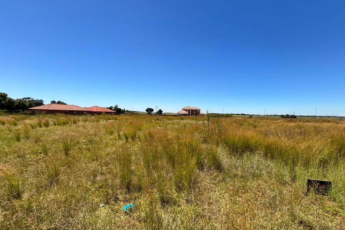 A wide-angle view of a vacant residential plot featuring natural grassland with surrounding neighborhood properties visible in the distance.