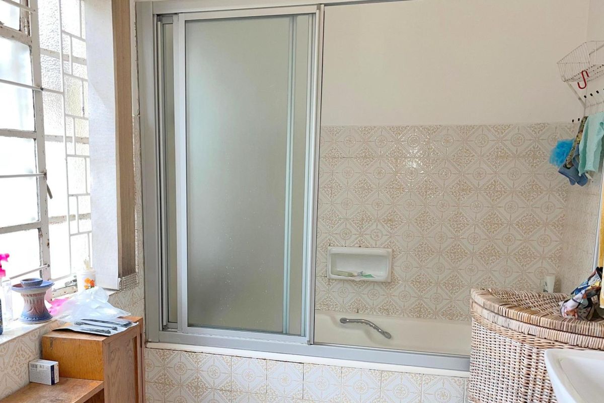 A dated bathroom featuring a built-in bathtub with a sliding glass shower screen and patterned ceramic wall tiles.
