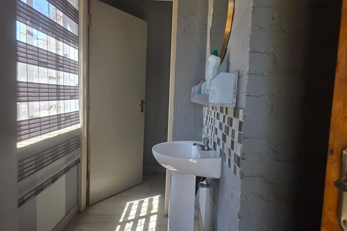 A compact bathroom featuring a pedestal basin, tiled splashback, and a window with modern blinds, leading into an adjacent room.