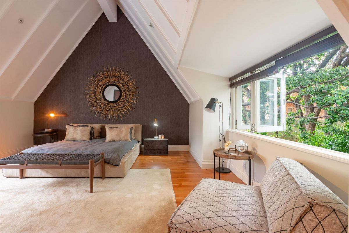 Spacious attic-style bedroom featuring high vaulted ceilings, exposed beams, hardwood flooring, and large windows overlooking mature trees.