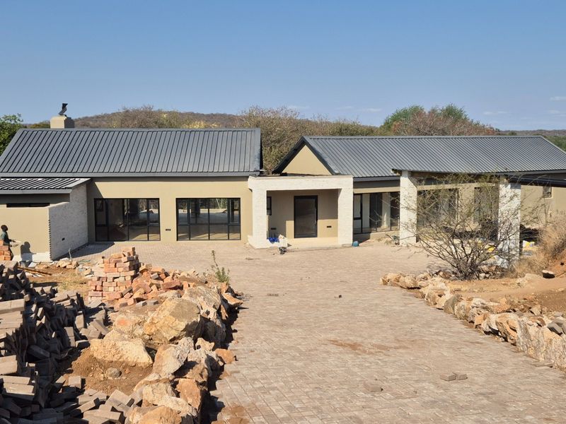 A modern single-story house under construction featuring a metal roof, stone accents, and a large paved driveway in a natural bushveld setting.