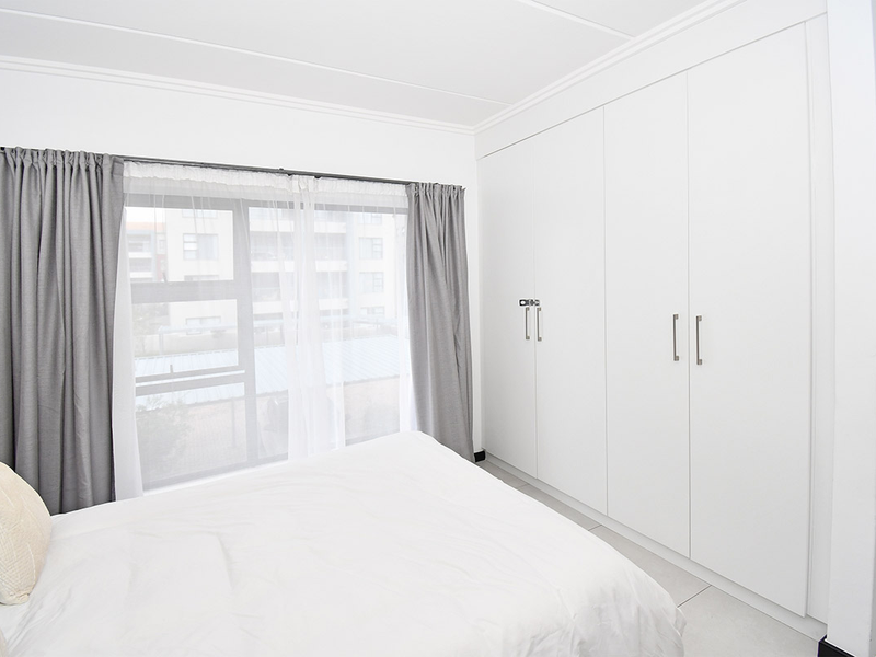 A bright bedroom featuring neutral tiled flooring, modern white built-in cupboards, and large aluminium-framed windows with curtain tracks.