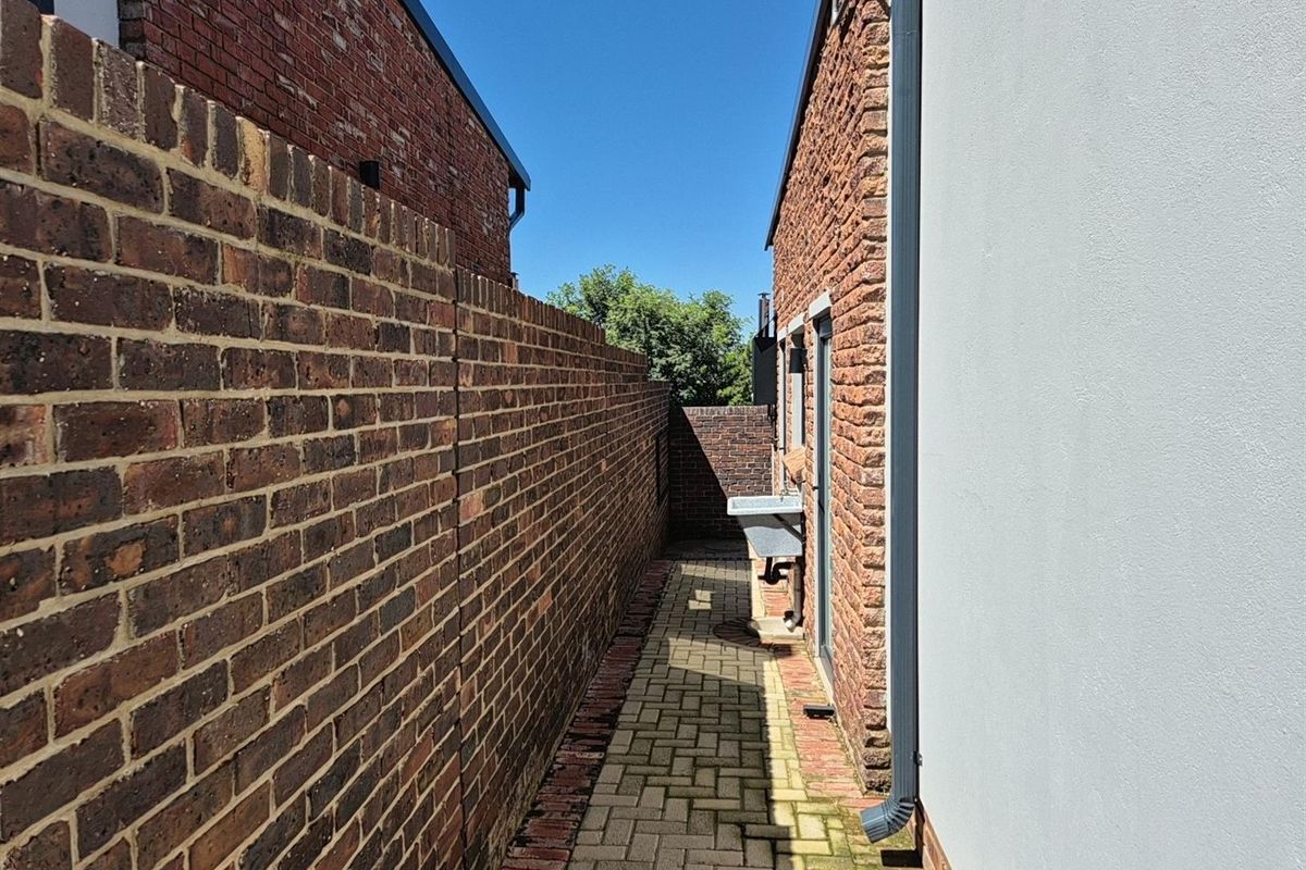 A narrow side passage featuring brick boundary walls, paved walkway, and a fixed outdoor utility sink attached to the building exterior.