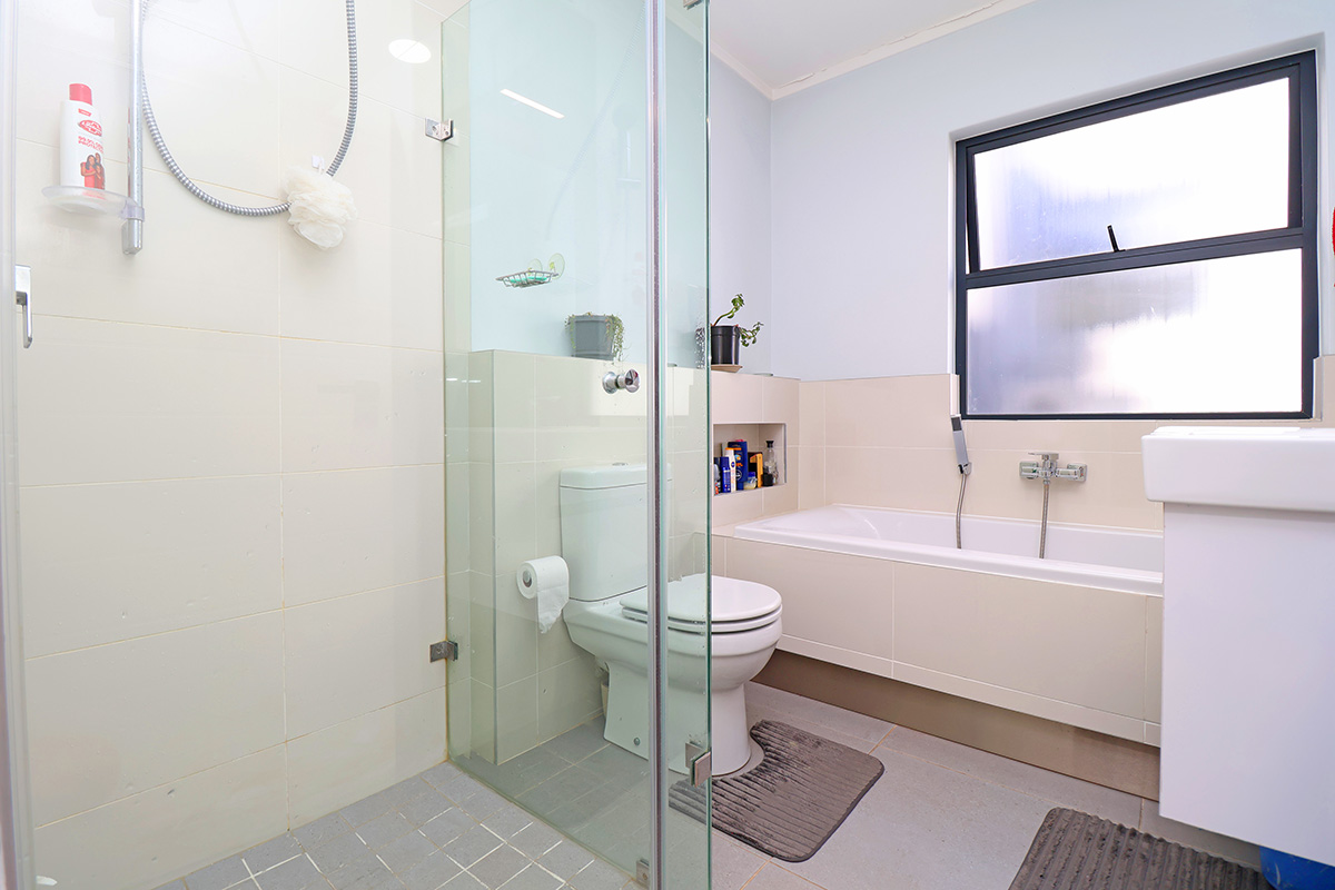 A functional bathroom featuring a glass-enclosed shower, a built-in bathtub, a toilet, and a vanity basin with neutral wall and floor tiling.