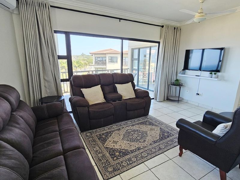 A bright and spacious living room featuring large sliding glass doors that open onto a balcony, tiled flooring, and a ceiling fan.