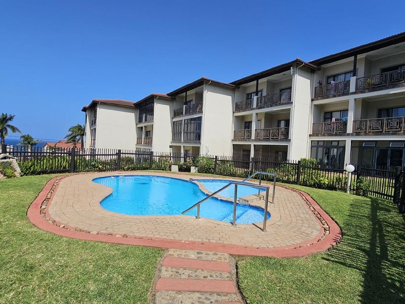 A communal swimming pool area set within a landscaped garden, featuring paved surrounds and views of the apartment complex exterior.