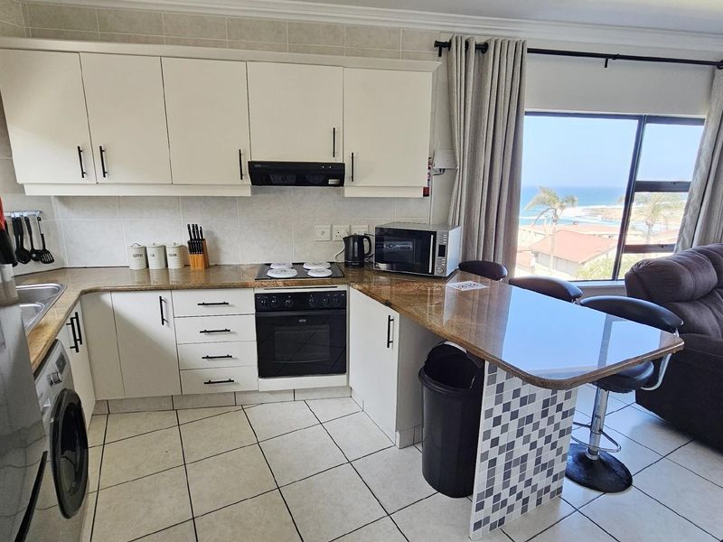 Open-plan kitchen featuring cream cabinetry, granite countertops, built-in oven and hob, and a breakfast bar, seamlessly flowing into the living area with ocean views.