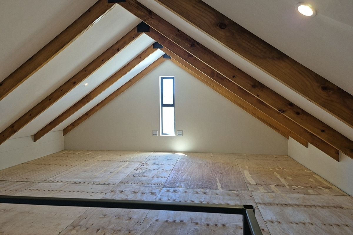 An attic-style loft space featuring exposed timber rafters, a central window, and unfinished plywood flooring, offering potential for a bedroom or home office.
