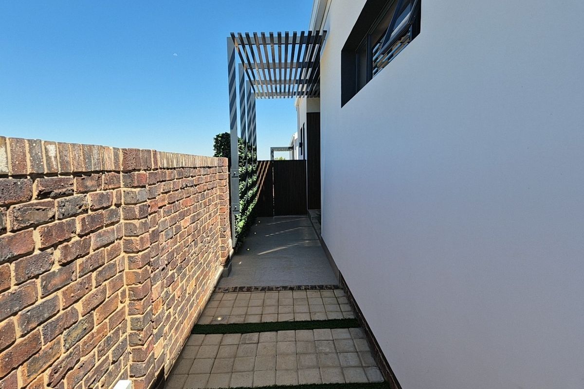 A side walkway featuring a brick boundary wall, paved path with grass inserts, and a modern overhead pergola structure.