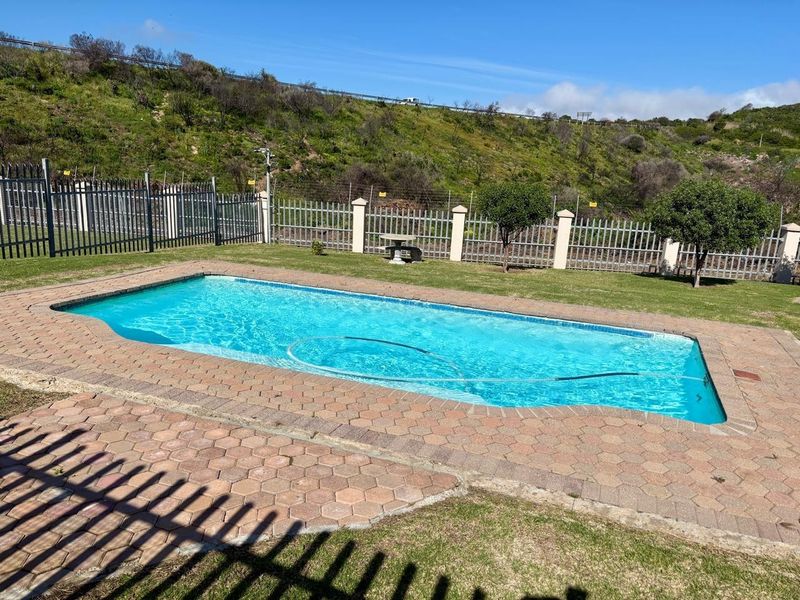 A well-maintained outdoor swimming pool area featuring paved surrounds, a manicured lawn, and secure metal perimeter fencing against a hillside backdrop.