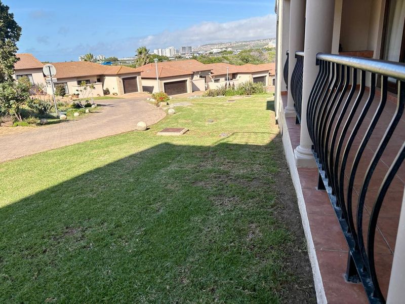 A covered patio area featuring a wrought iron balustrade and tiled flooring, overlooking a residential townhouse complex and landscaped garden.