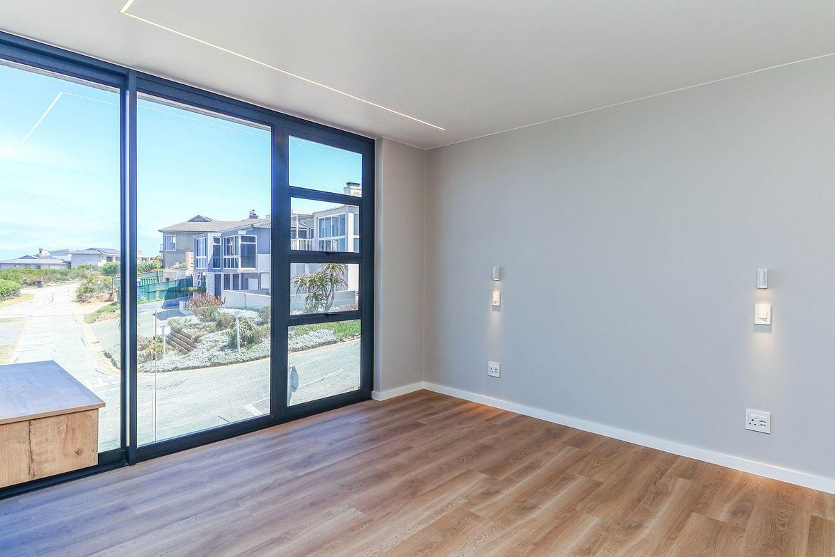 A bright, modern bedroom featuring high-quality wood-look flooring, recessed ceiling lighting, and large floor-to-ceiling sliding glass doors with views of the neighborhood.