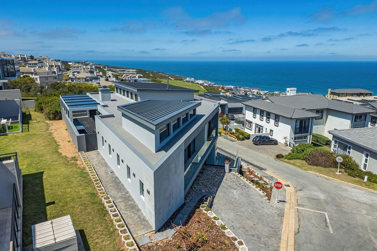 Aerial view of a modern coastal residence featuring a contemporary flat roof design, paved driveway, and landscaped surroundings with ocean views.