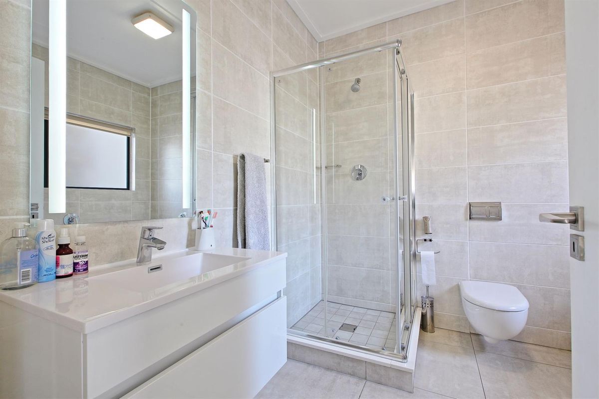 Modern bathroom featuring a wall-hung toilet, glass shower enclosure, and a sleek vanity with an illuminated mirror and neutral floor-to-ceiling tiling.