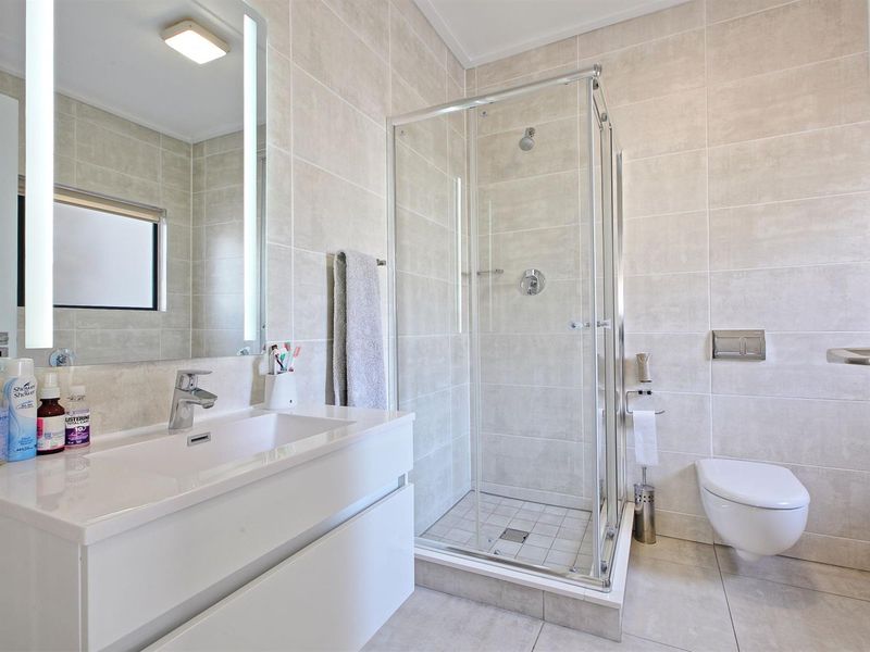 Modern bathroom featuring a wall-hung toilet, glass shower enclosure, and a sleek vanity with an illuminated mirror and neutral floor-to-ceiling tiling.