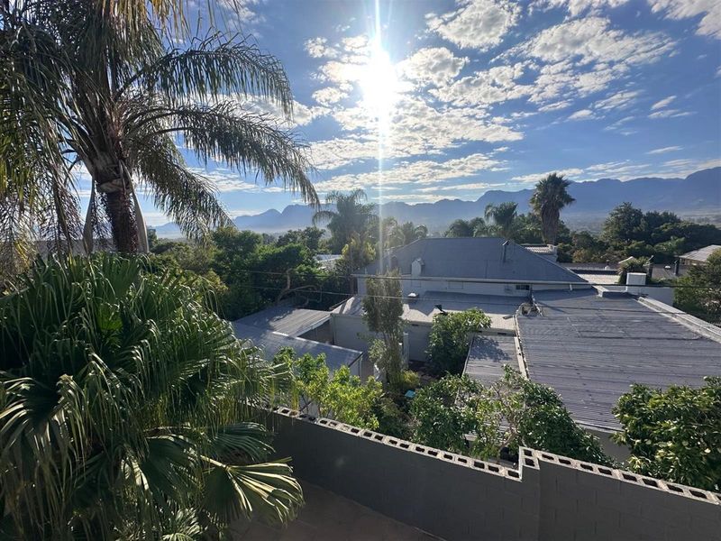 Elevated view overlooking the property roofline, surrounding garden, and distant mountain range under a bright sky.