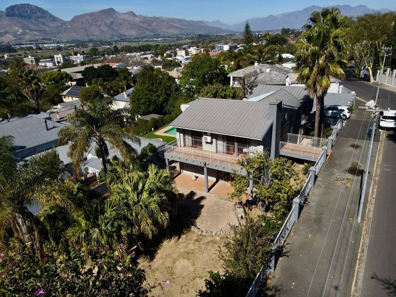 Aerial view of a two-story residential property featuring a metal roof, upper-level balcony, and a paved patio area set within a landscaped garden.