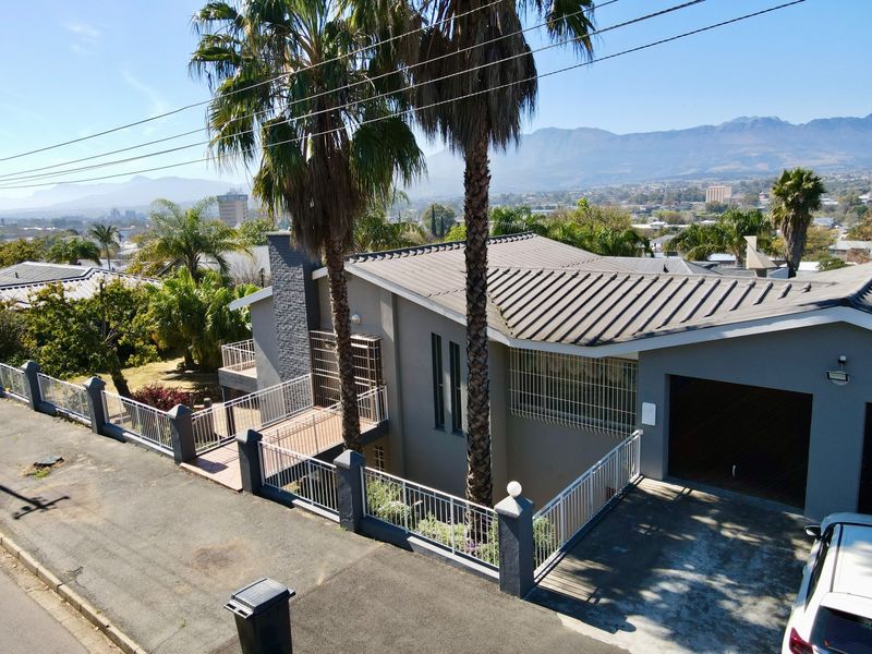 Aerial view of a modern house featuring a metal roof, secure perimeter fencing, and a paved driveway with mountain views in the background.