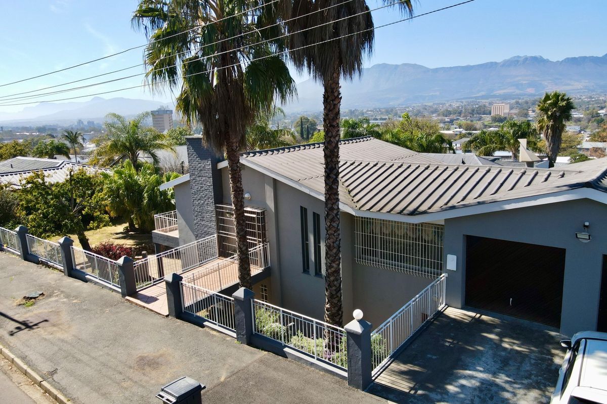 Aerial view of a modern house featuring a metal roof, secure perimeter fencing, and a paved driveway with mountain views in the background.