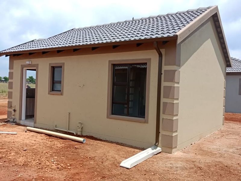 A newly constructed single-story house featuring a tiled roof, aluminium window frames, and exterior plumbing connections, set on an undeveloped plot.