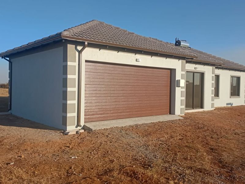 Exterior view of a modern single-story house featuring a tiled roof, integrated garage, and solar water heating system, set on an undeveloped plot.