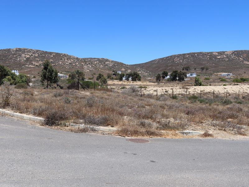 A street-level view of a vacant residential plot featuring asphalt road access, concrete curbing, and perimeter wire fencing against a hilly landscape.